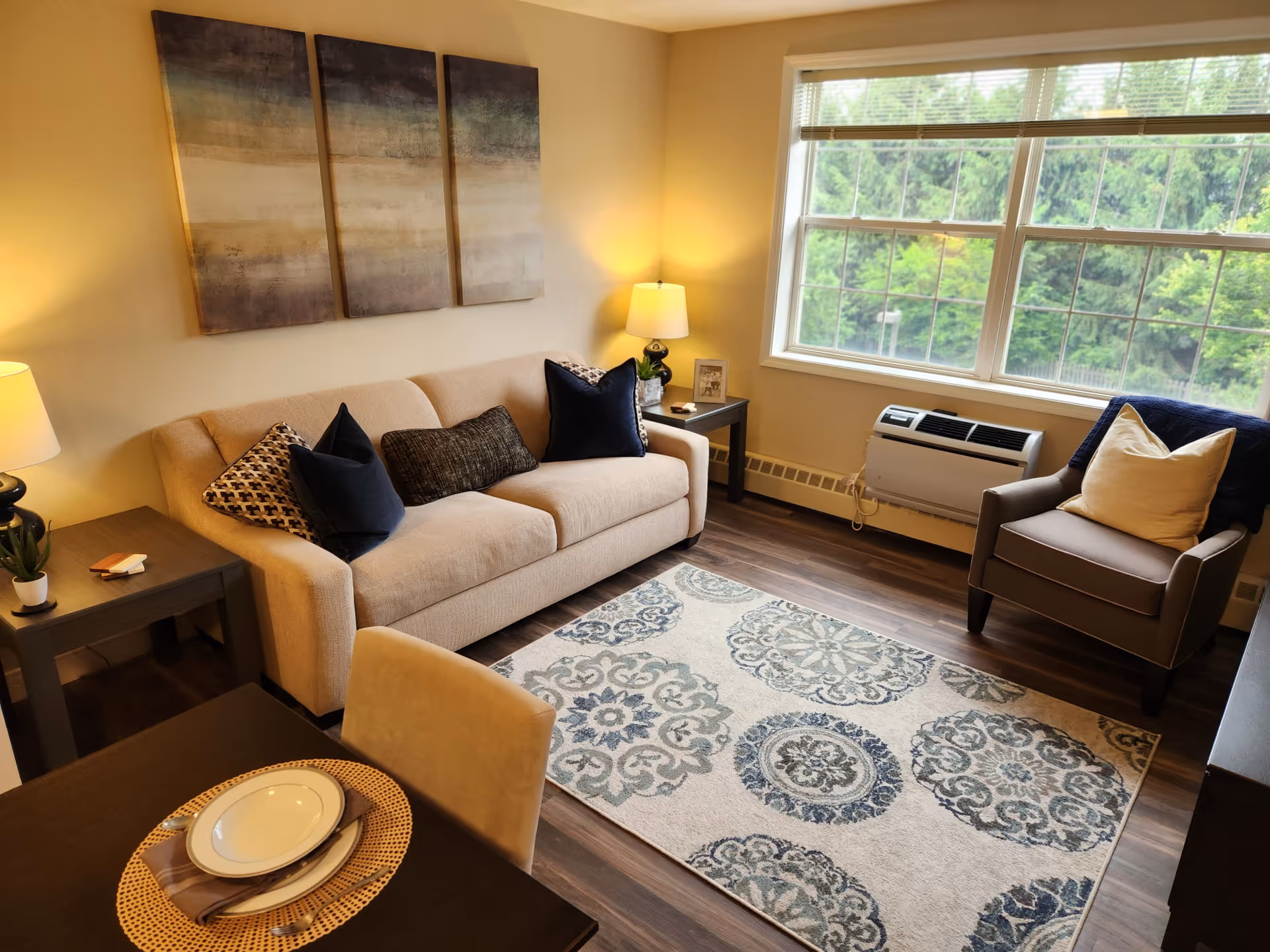 A cozy living room with a beige sofa adorned with multiple pillows, a patterned area rug on wooden flooring, a single armchair with a pillow and throw blanket, two side tables with lamps, and a large window showing green trees outside. There is also a small dining table with a place setting in the foreground.