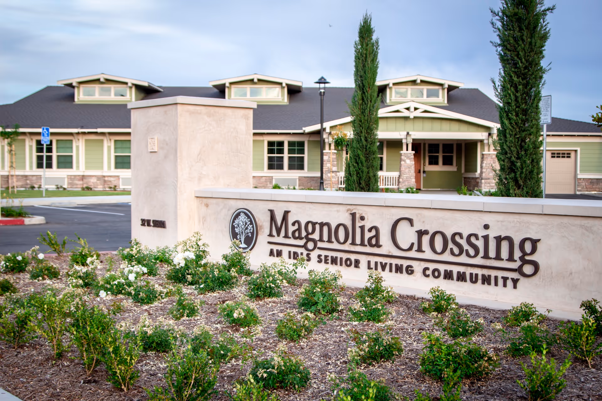 Entrance sign for Magnolia Crossing, an IDES senior living community, with a landscaped garden in front and a large building with green siding and a dark roof in the background.