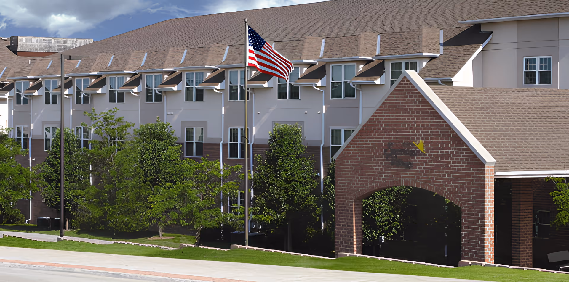 Exterior view of a multi-story retirement community building with beige and brick facade, multiple windows, and a large American flag on a flagpole in front. There are green trees and a well-maintained lawn surrounding the building under a partly cloudy sky.