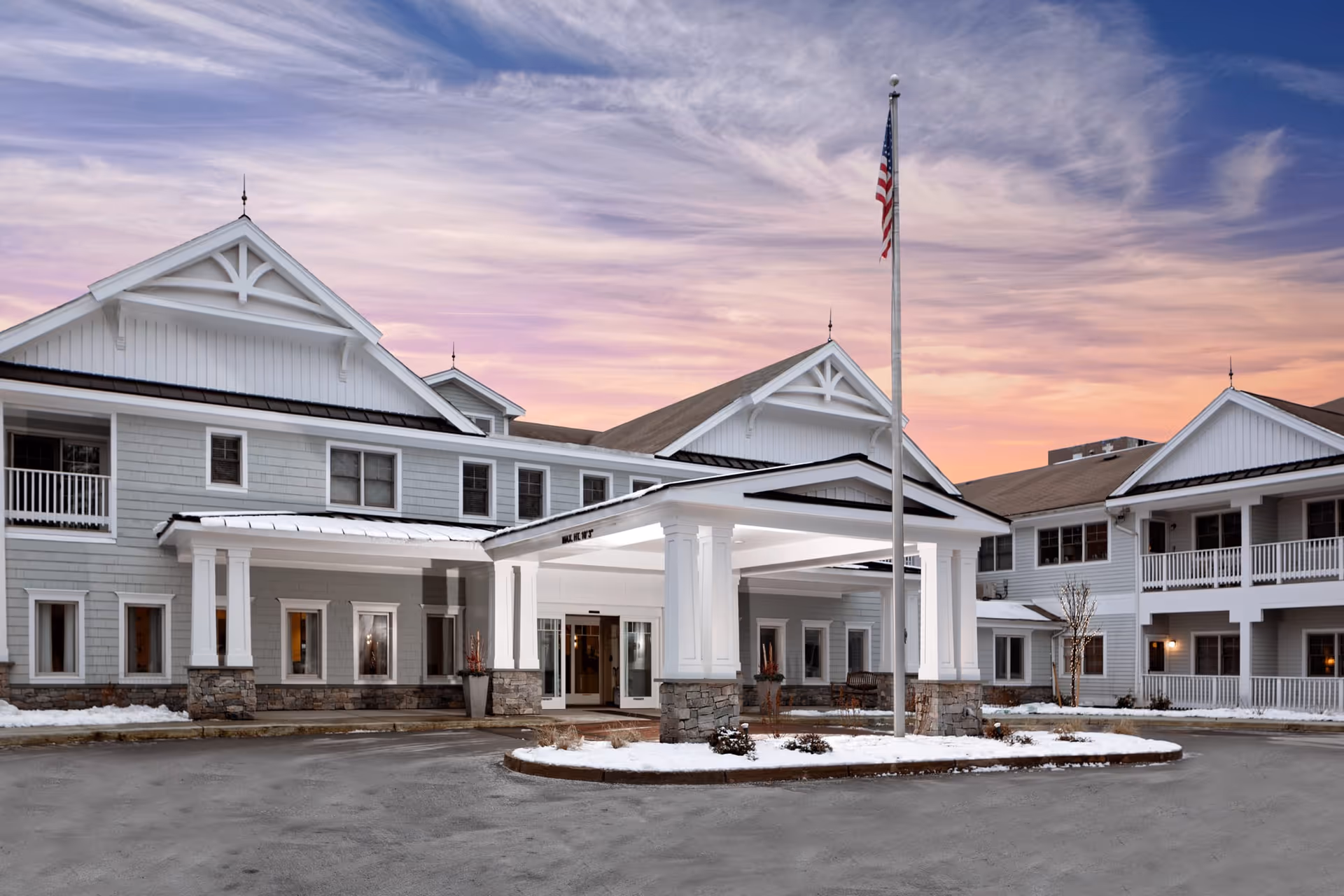 Exterior view of a senior living facility building named Maplewood at Weston during sunset, featuring a covered entrance with white pillars, stone accents, and an American flag on a flagpole in front. There is some snow on the ground around the building.