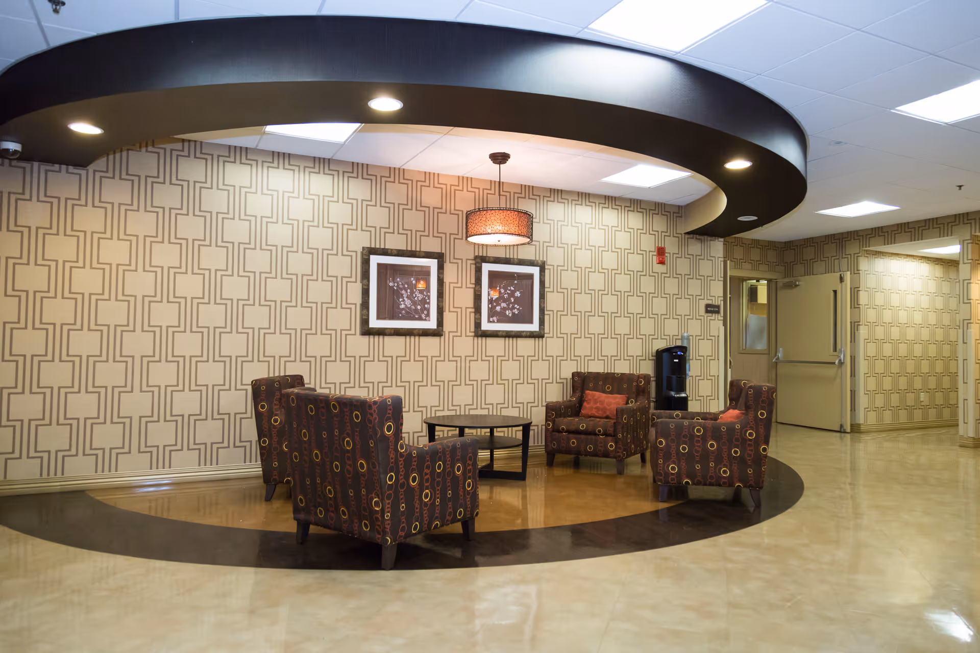 A seating area in a senior living facility with four patterned armchairs arranged around a small round table. The walls have a geometric patterned wallpaper, and two framed pictures hang on the wall. A circular ceiling feature with recessed lighting is above the seating area. There is a water dispenser and double doors in the background.