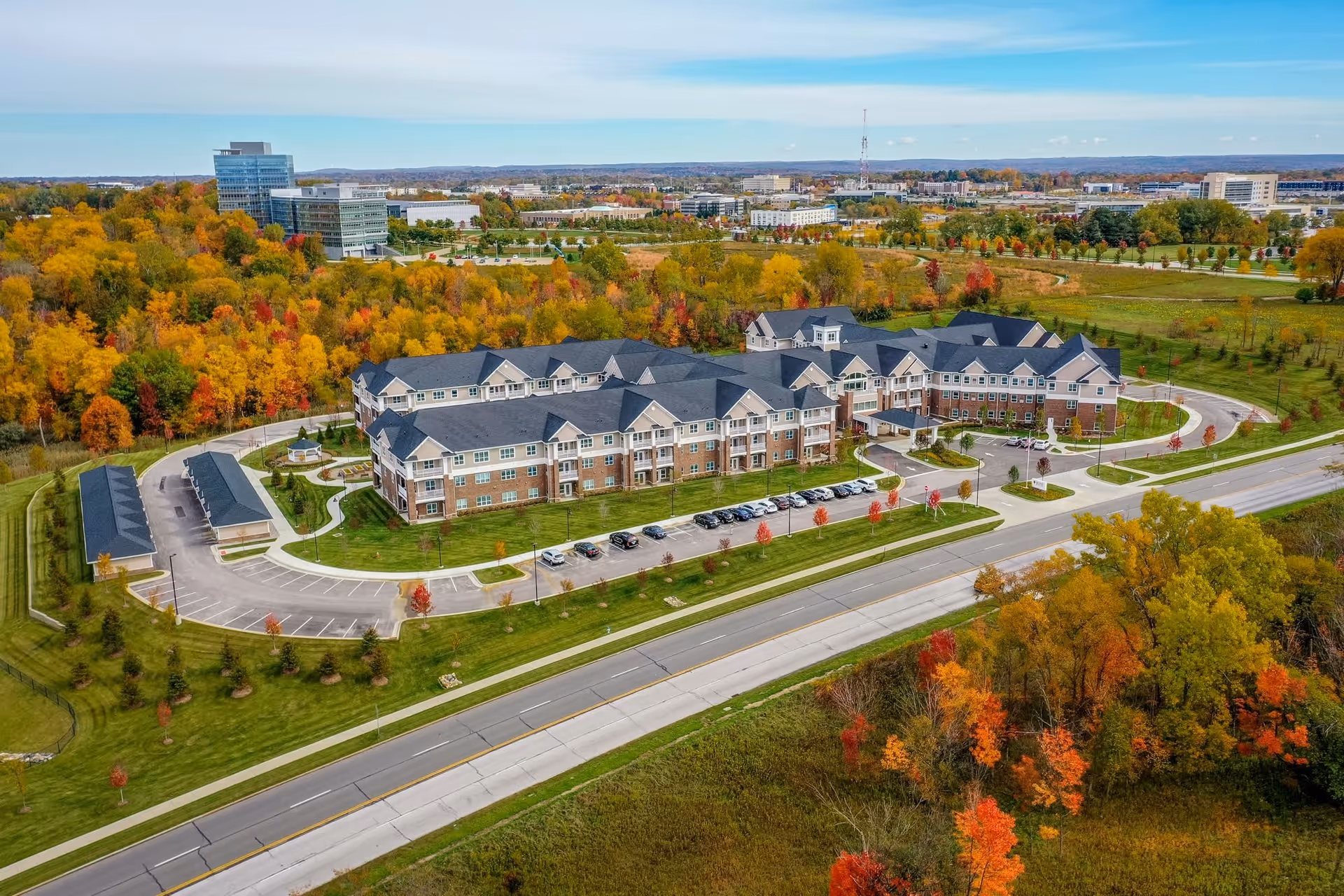 Aerial view of Rose Senior Living Beachwood, a large senior living facility surrounded by autumn trees with colorful foliage. The building has a multi-wing design with a dark roof and brick and light-colored exterior walls. There are parking lots with cars and a road in the foreground, with a backdrop of office buildings and open green spaces.