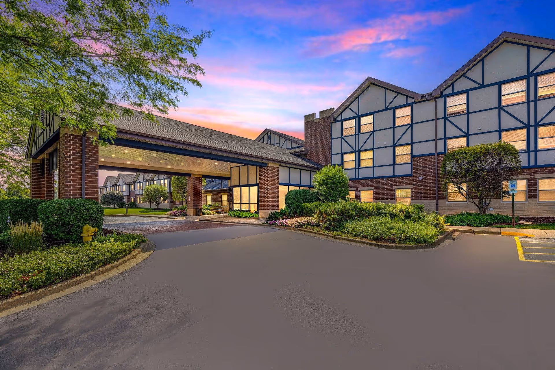 Exterior view of Sunrise of Olympia Fields senior living facility at sunset, showing a large covered entrance with brick pillars, landscaped greenery, and a multi-story building with illuminated windows under a colorful sky.