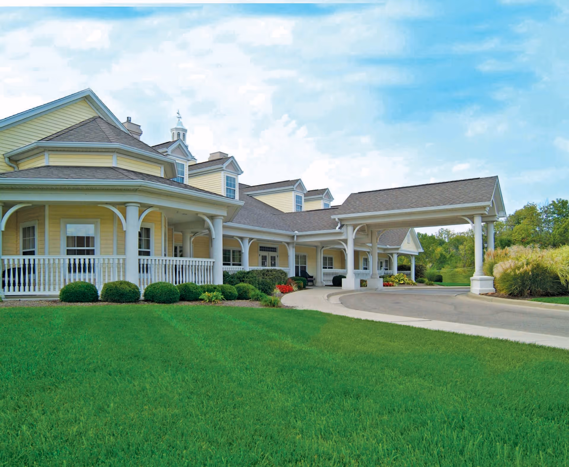 Exterior view of a senior living facility with yellow siding, white columns, and a covered driveway entrance. The building is surrounded by a well-maintained green lawn and some shrubs, under a partly cloudy blue sky.