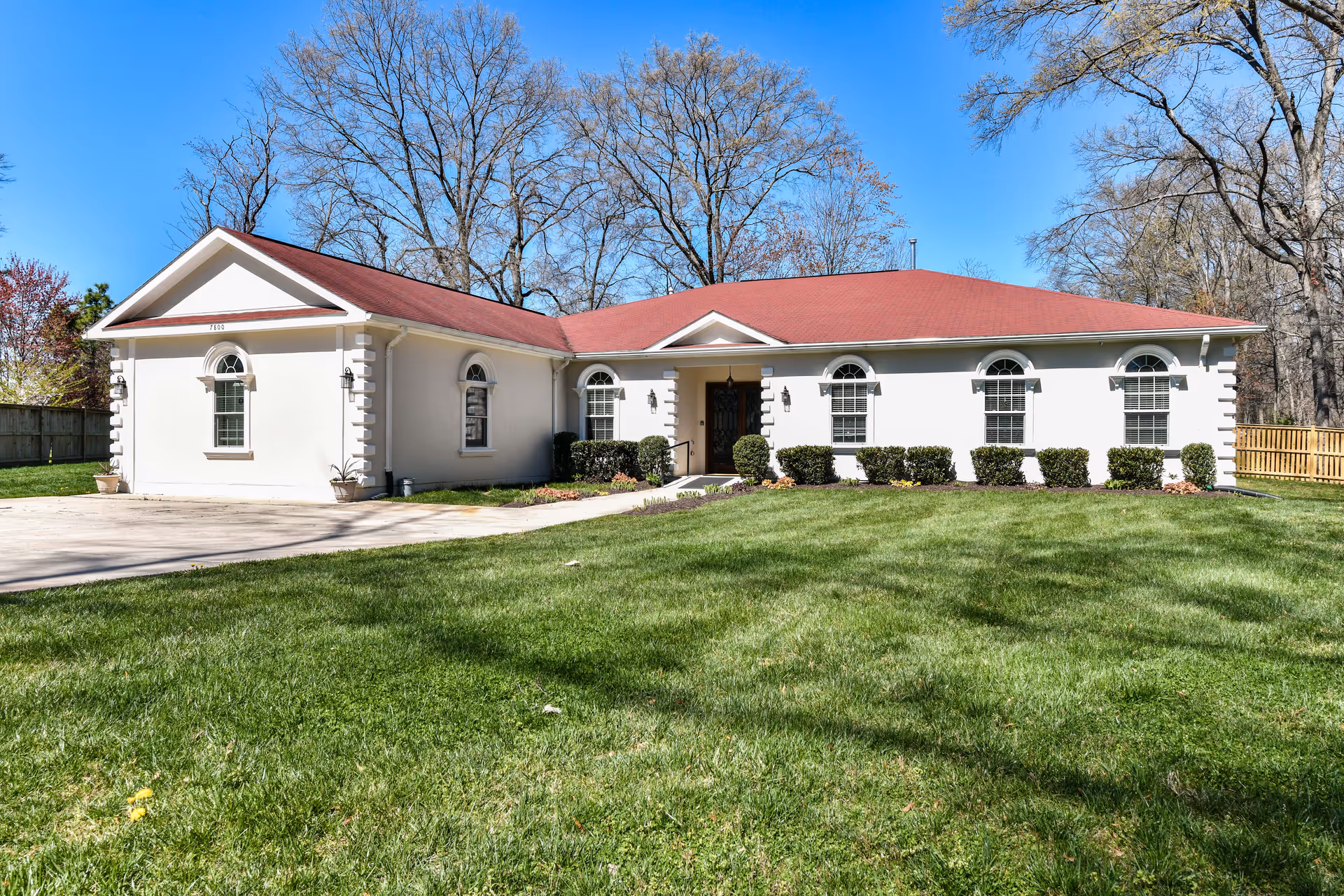 Front exterior view of a single-story building with a red roof, white walls, and multiple arched windows. The building is surrounded by a well-maintained green lawn and some bushes near the entrance. Trees without leaves are visible in the background under a clear blue sky.