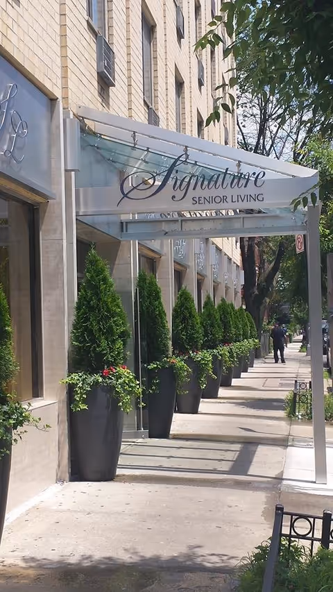 Sidewalk view of the exterior entrance to Signature Senior Living facility with a row of tall potted plants along the building and a glass canopy with the facility's name above the entrance.