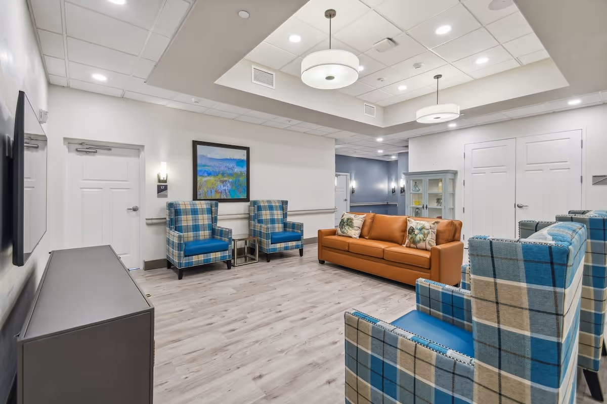 A bright and modern living room area in a senior living facility featuring a brown leather sofa with floral pillows, four blue and beige plaid armchairs, light wood flooring, a wall-mounted TV, a colorful landscape painting, and soft ceiling lighting fixtures.