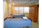 A rehabilitation room with two hospital-style beds covered in blue sheets, positioned parallel to each other. The room has a large window with a beige valance, letting in natural light. There is a wooden cabinet with multiple drawers and doors on the right side of the room. The walls are painted light yellow and beige, and the floor is covered with light-colored tiles.