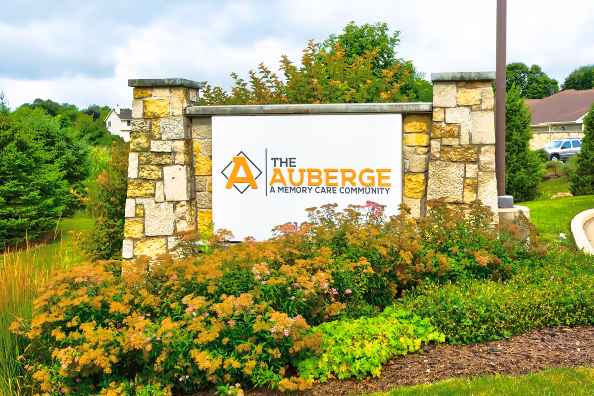 Stone sign for The Auberge, a memory care community, surrounded by green shrubs and flowering plants with a partly cloudy sky in the background.