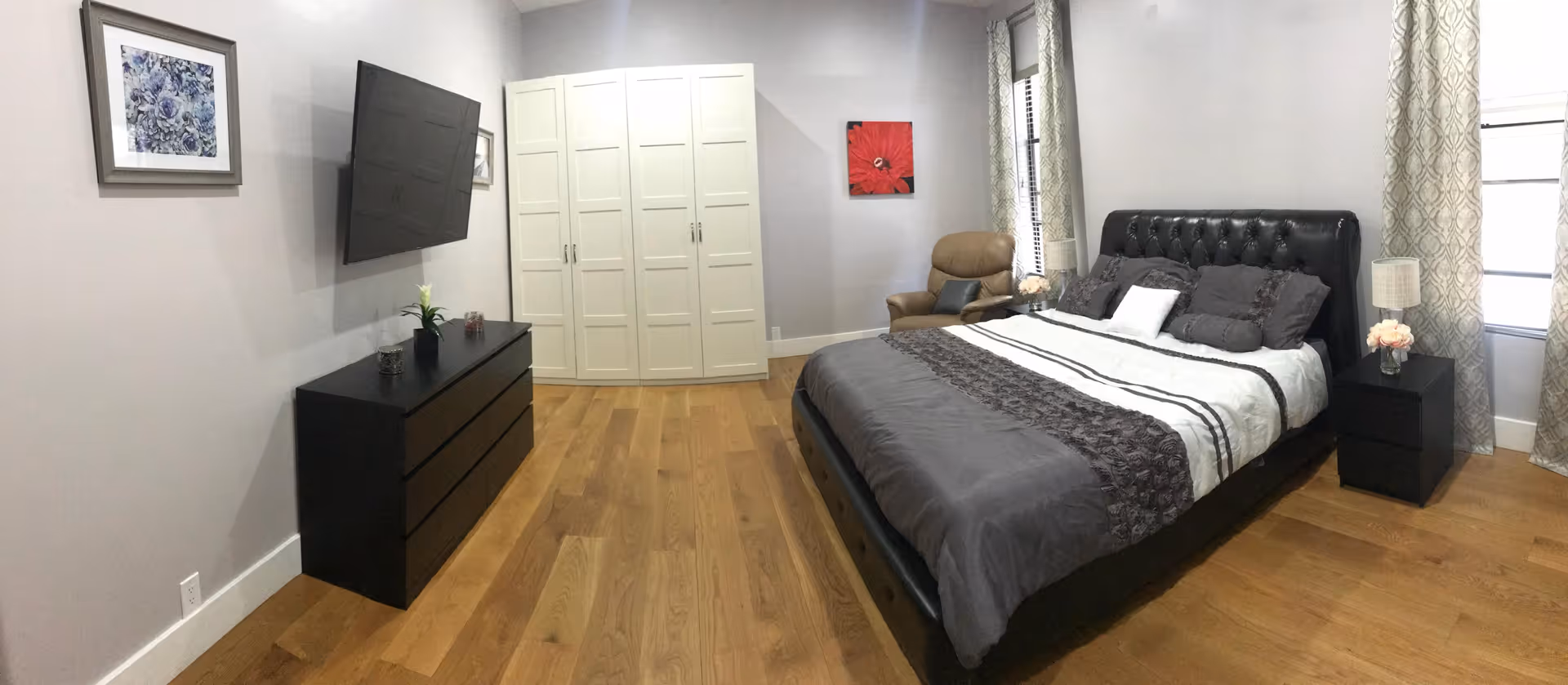 A modern bedroom with a large bed featuring gray and white bedding, a black tufted headboard, and multiple pillows. To the right of the bed is a black nightstand with a lamp and a small vase of flowers. A brown armchair is positioned near two windows with patterned curtains. On the opposite wall, there is a black dresser with decorative items and a flat-screen TV mounted above it. A white wardrobe is placed against the back wall, and the room has wooden flooring and light gray walls with framed artwork.
