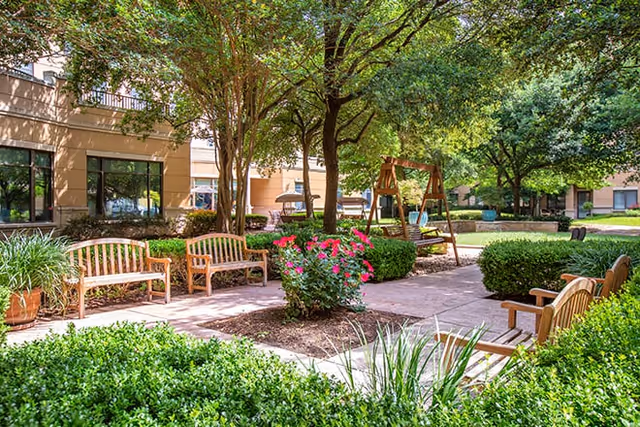 Outdoor garden area at Brookdale Gaines Ranch featuring wooden benches, a swing set, green bushes, flowering plants, and trees providing shade near the building exterior.