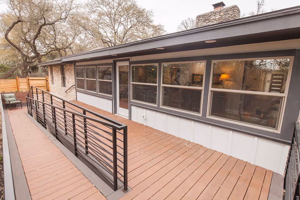 Exterior view of a senior living facility showing a wooden deck with a black metal railing and a ramp leading to a door. The building has large windows and a stone wall section, with trees and a wooden fence in the background.