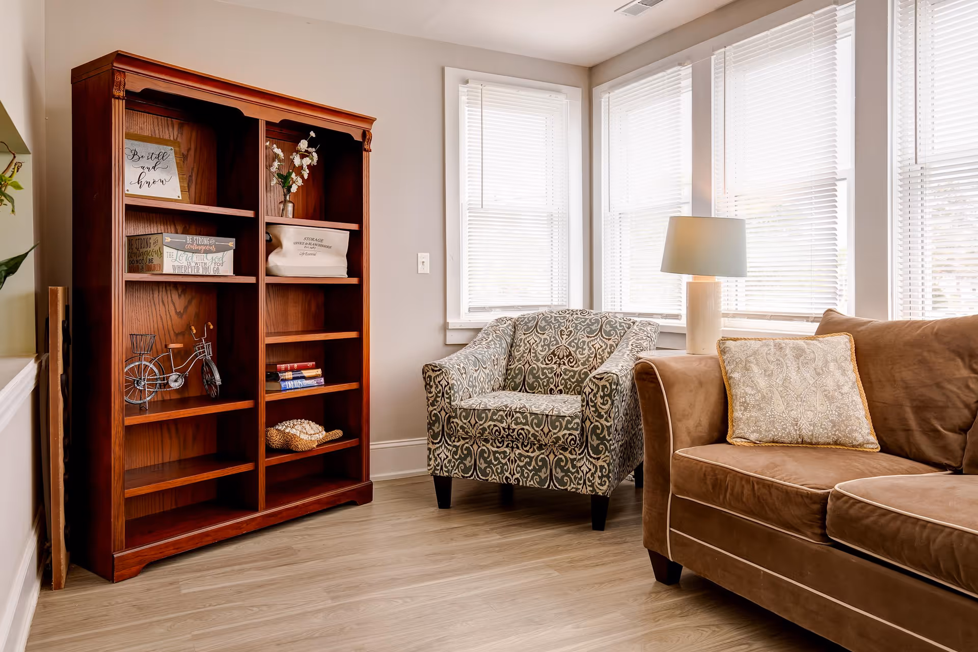 A cozy living room area with a patterned armchair, a brown sofa with a decorative pillow, a wooden bookshelf with various decorative items and books, and a table lamp near windows with white blinds letting in natural light.