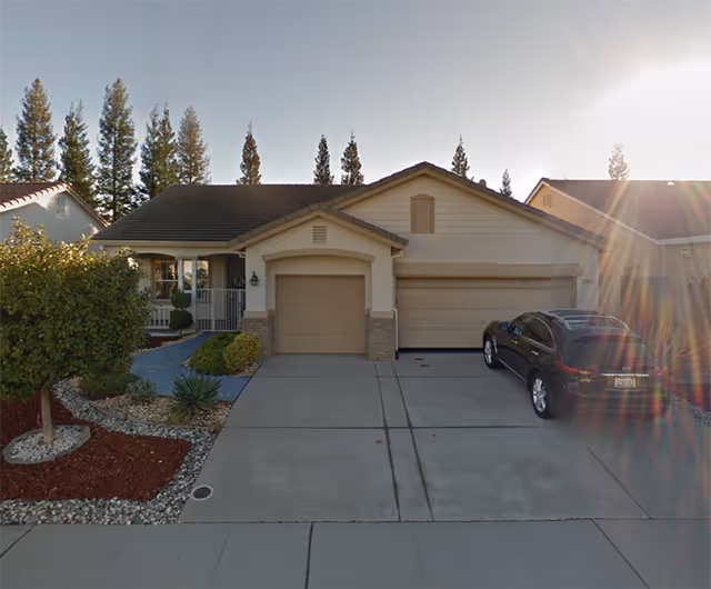 Front exterior view of a single-story house with a two-car garage and a driveway. There is a black car parked on the right side of the driveway. The house has a small front porch with a white railing and some landscaping including a tree and bushes. Tall pine trees are visible in the background under a clear sky with sunlight creating lens flare.