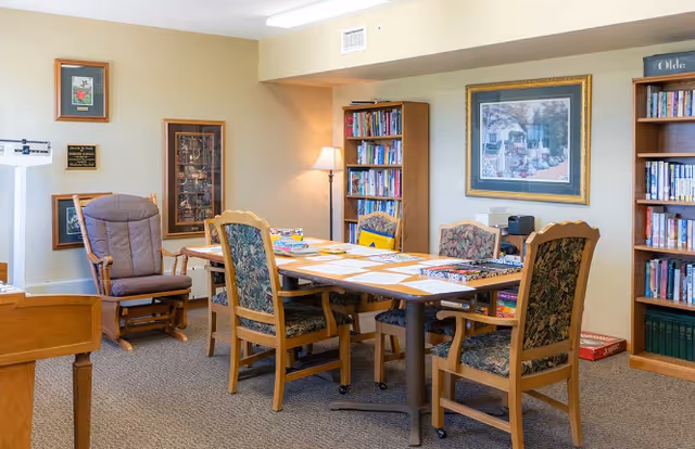 A cozy interior room with a rectangular table surrounded by six wooden chairs with patterned cushions. The table has papers and board games on it. There is a cushioned rocking chair to the left, two tall bookshelves filled with books, a floor lamp, and framed artwork on the walls. The room has beige walls and carpeted flooring.