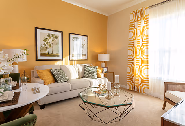 A bright and cozy living room with a beige sofa adorned with green and yellow patterned cushions, a glass coffee table with decorative candles and geometric sculpture, a round dining table set with plates and glasses, two framed botanical prints on a warm yellow wall, a window with sheer white curtains and yellow patterned drapes, and a side table with a lamp and picture frame.