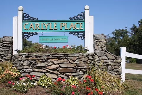 Stone sign with white posts and decorative black metal accents reading 'CARLYLE PLACE' with a smaller green sign below stating 'A COURVILLE COMMUNITY', surrounded by flowers and greenery under a clear blue sky.