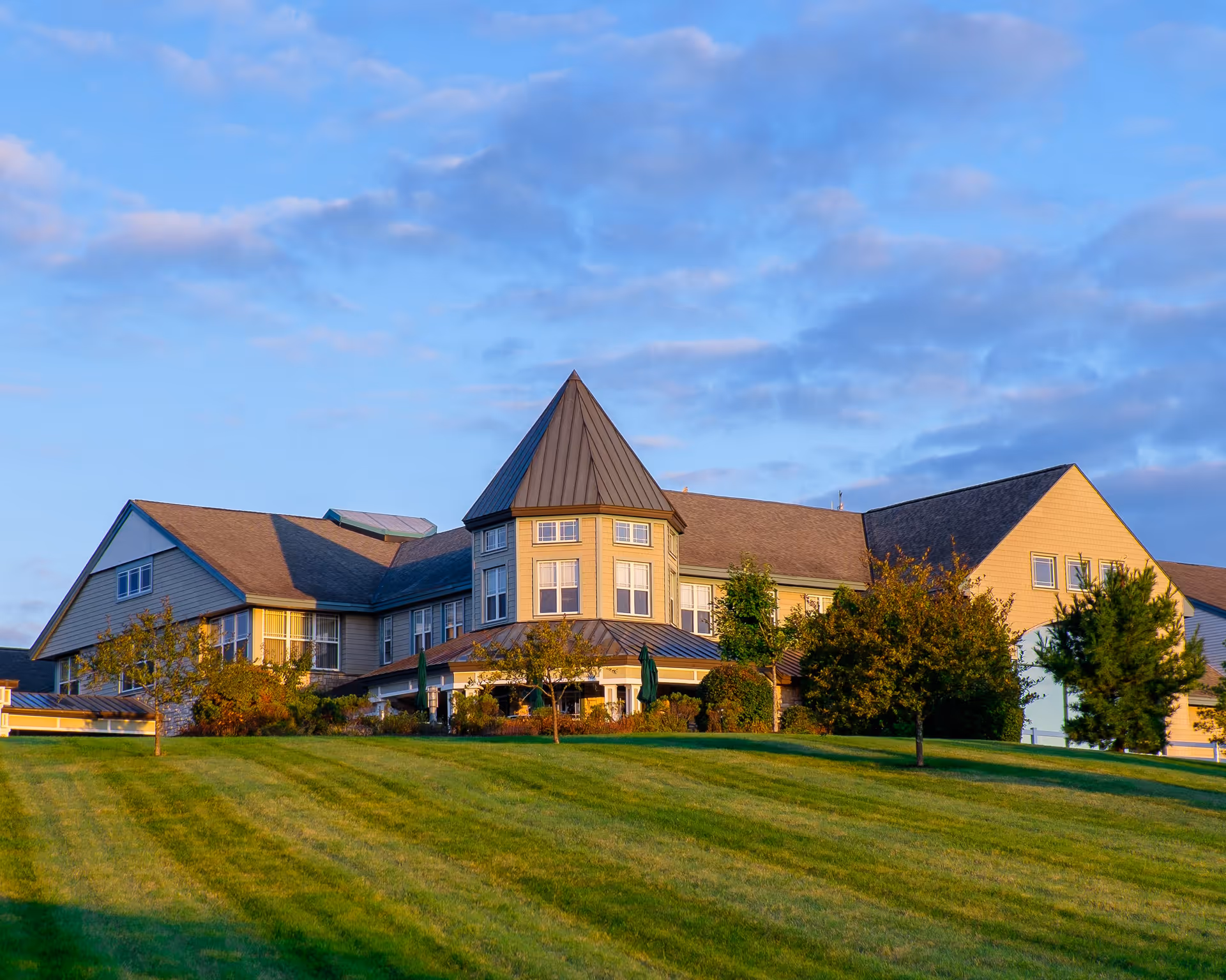 Exterior view of a large senior living facility building named Glen Eddy, featuring a distinctive turret and surrounded by a well-maintained grassy lawn with trees under a partly cloudy sky during sunset.