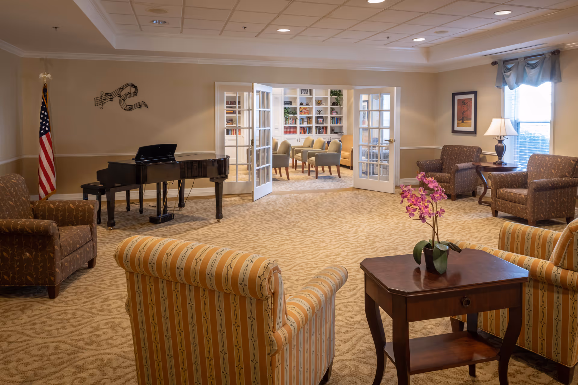 A spacious senior living common area with patterned carpet, several upholstered armchairs and sofas, a wooden side table with a potted orchid, a black grand piano near an American flag, and French doors leading to a bright room with bookshelves and additional seating.