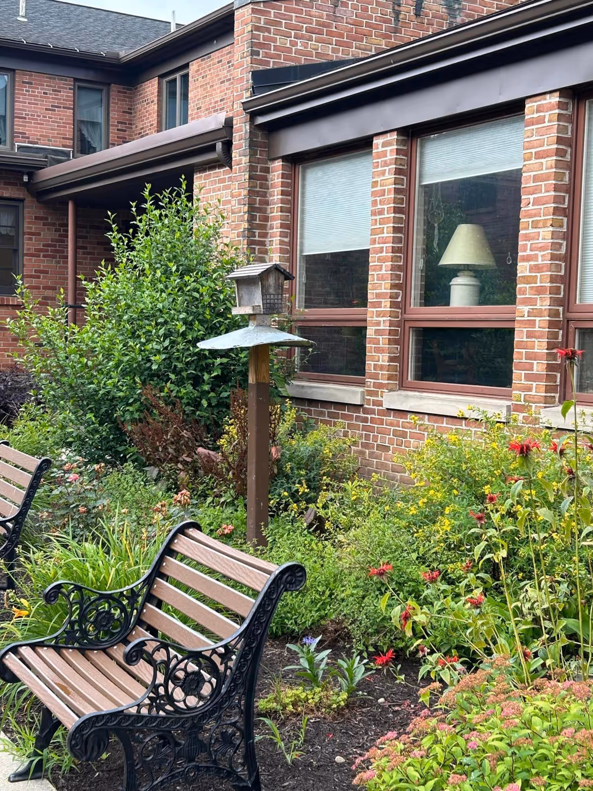 Outdoor garden area with two wooden benches featuring ornate black metal armrests and legs, surrounded by various green plants and colorful flowers. A birdhouse on a post is visible among the plants, and a brick building with large windows and a lamp inside is in the background.