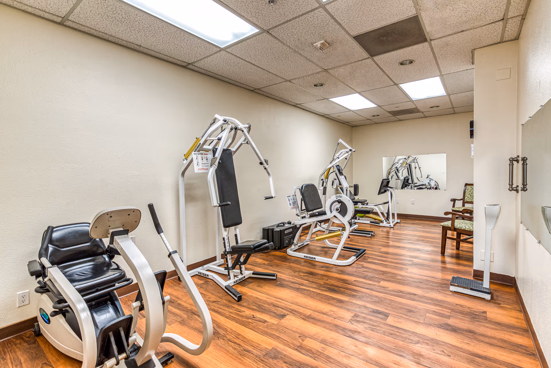 A small fitness room with wooden flooring and beige walls, equipped with several exercise machines including a recumbent bike and weight machines. There is a large mirror on the far wall and a wooden chair with green patterned upholstery near the corner.