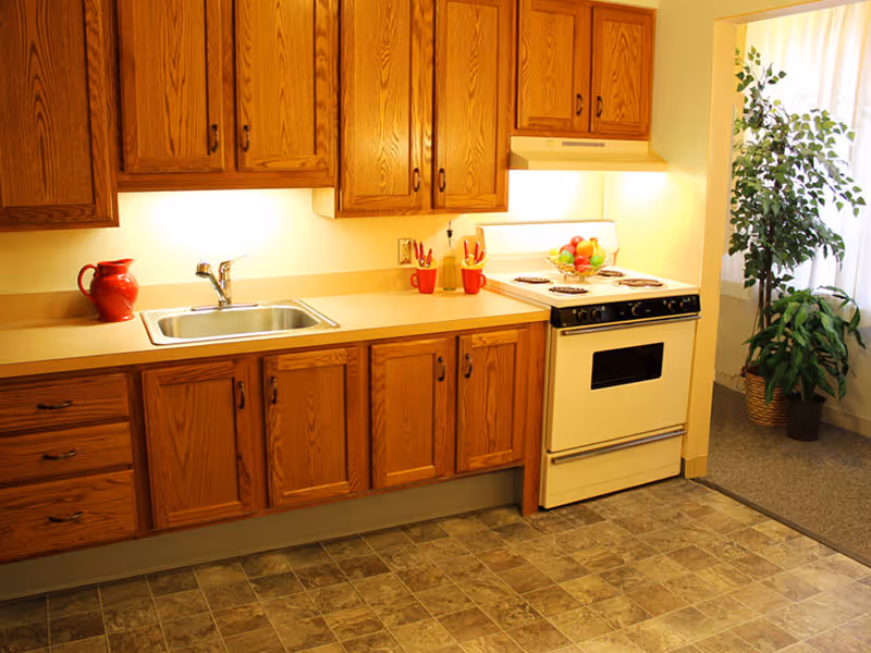 A kitchen area with wooden cabinets, a countertop with a sink, a red pitcher, and a stove with a fruit bowl on top. There is a potted plant near a window with sheer curtains in the adjacent room.