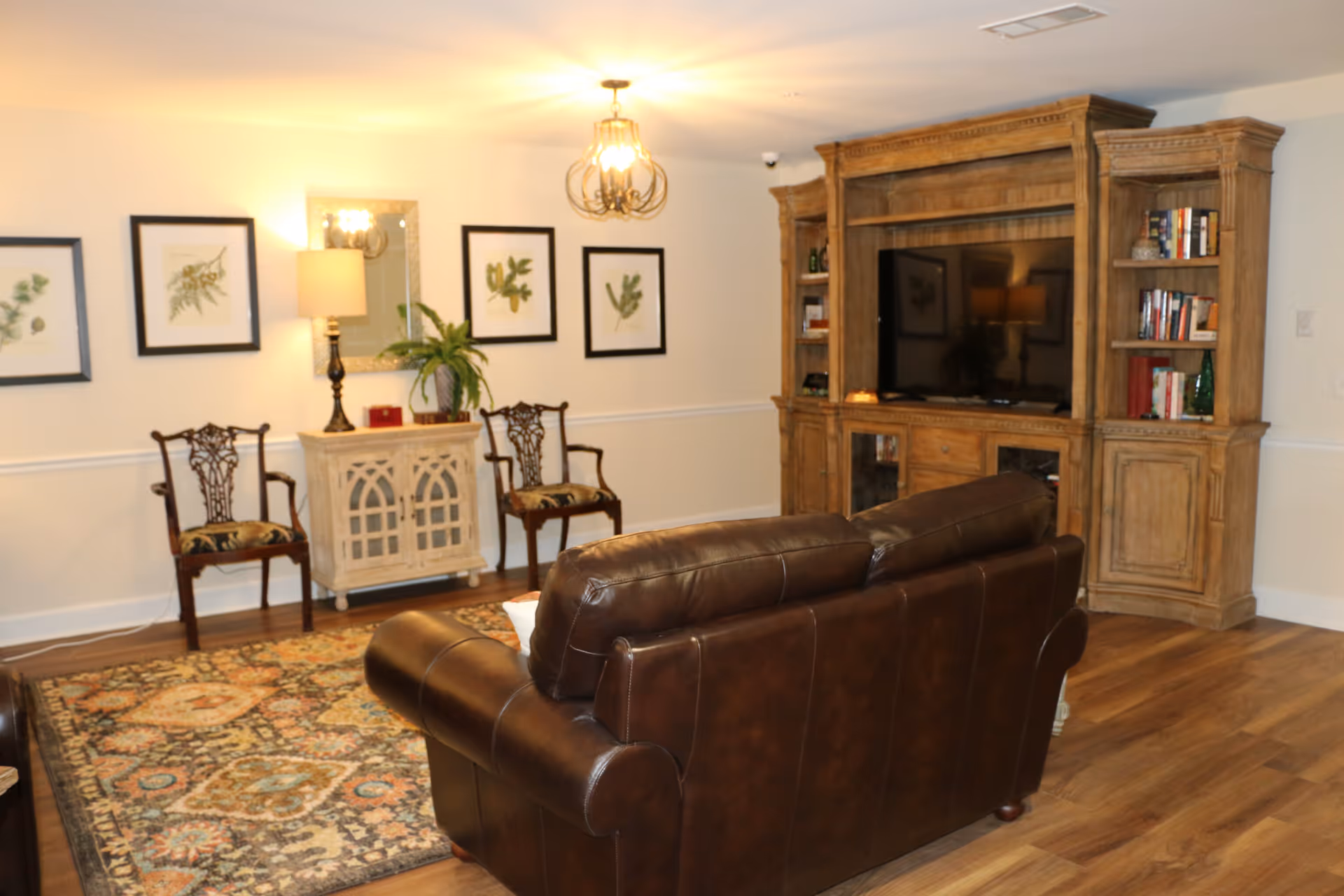A cozy living room with a brown leather sofa facing a large wooden entertainment center with a flat-screen TV. The room has wooden flooring, a patterned area rug, two wooden chairs with patterned cushions, a small cabinet with a lamp and a potted plant, and framed botanical prints on the wall.