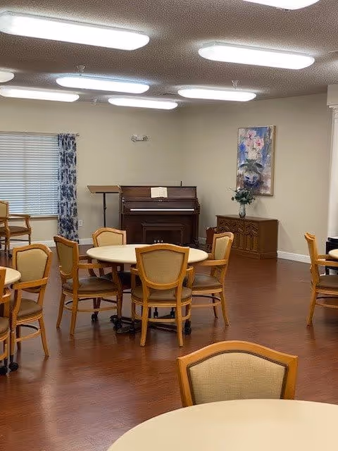 A well-lit room with round tables and wooden chairs arranged around them. There is a piano against the far wall with an open book on the music stand. A floral painting hangs on the wall above a wooden cabinet with a vase of flowers. The room has hardwood floors and a window with blue patterned curtains.