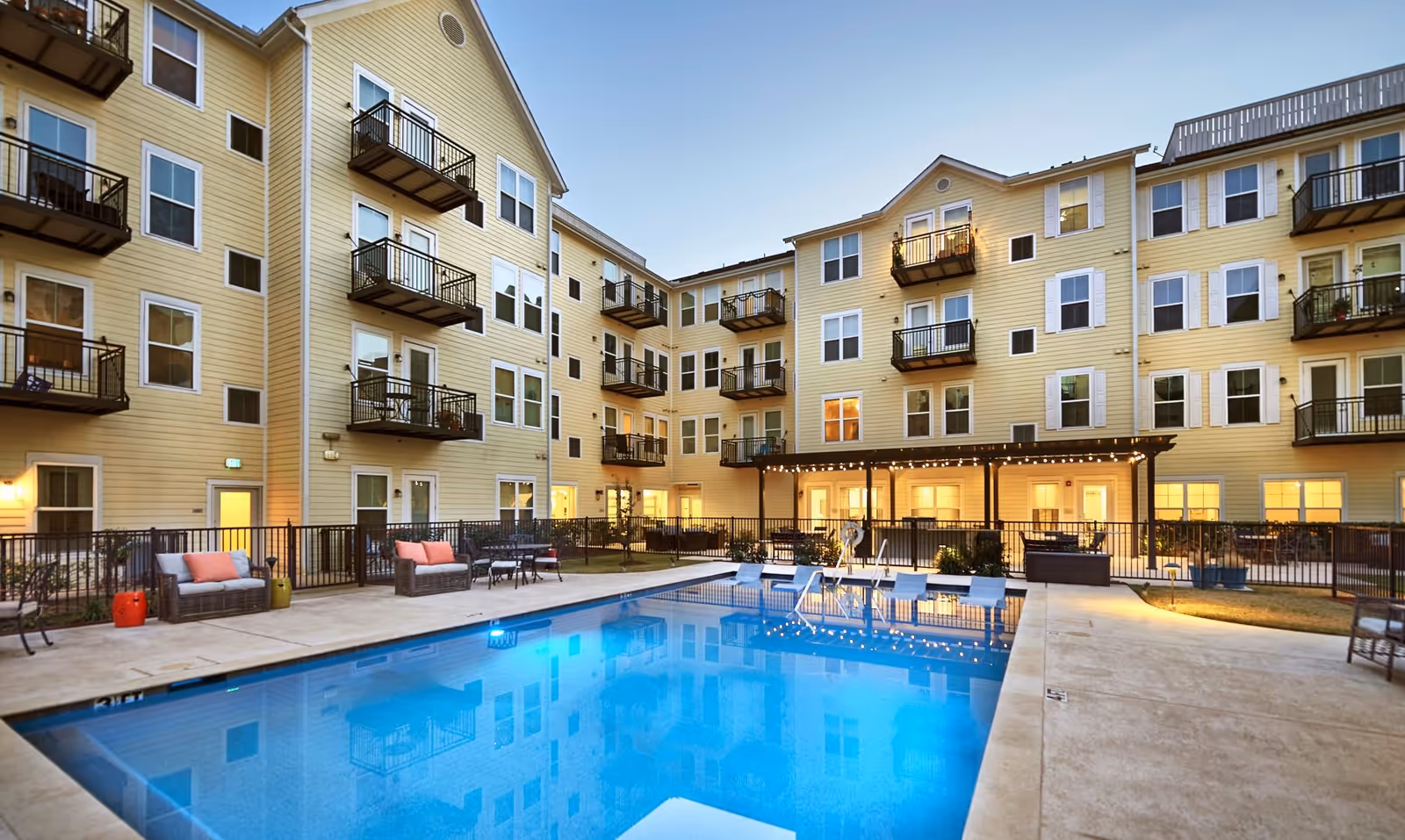 Outdoor view of a multi-story retirement facility with beige siding and multiple balconies surrounding a large swimming pool. The pool area has lounge chairs, seating areas with cushions, and is lit with string lights under a covered patio. The sky is clear and it appears to be early evening.