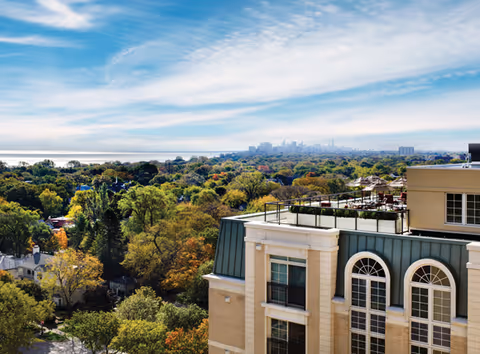 View from a high vantage point showing the rooftop and upper floors of a beige building with green roofing, surrounded by a dense area of trees with autumn foliage. In the distance, a city skyline is visible under a partly cloudy blue sky.
