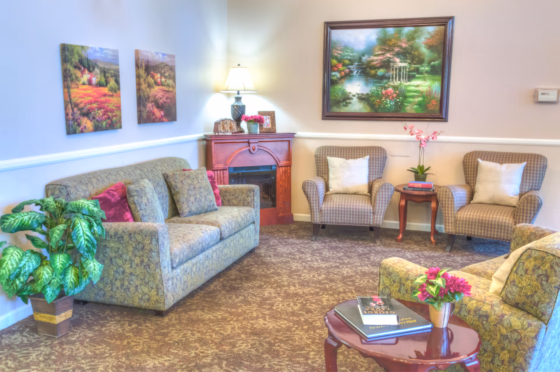 A cozy living room with two patterned armchairs and a matching sofa, all with cushions. There is a wooden side table with a lamp and a small plant, and a coffee table with books and a flower pot. The walls are decorated with colorful landscape paintings, and there is a large leafy plant in the corner.