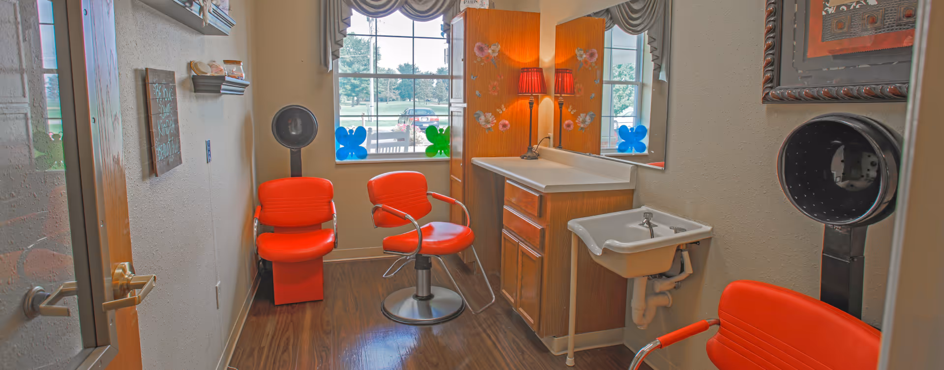 Interior view of a small salon or hair styling room with three bright red salon chairs, a wooden cabinet with a countertop and sink, a wall-mounted hair dryer, and a window decorated with colorful butterfly decals. The room has wood flooring and light-colored walls.