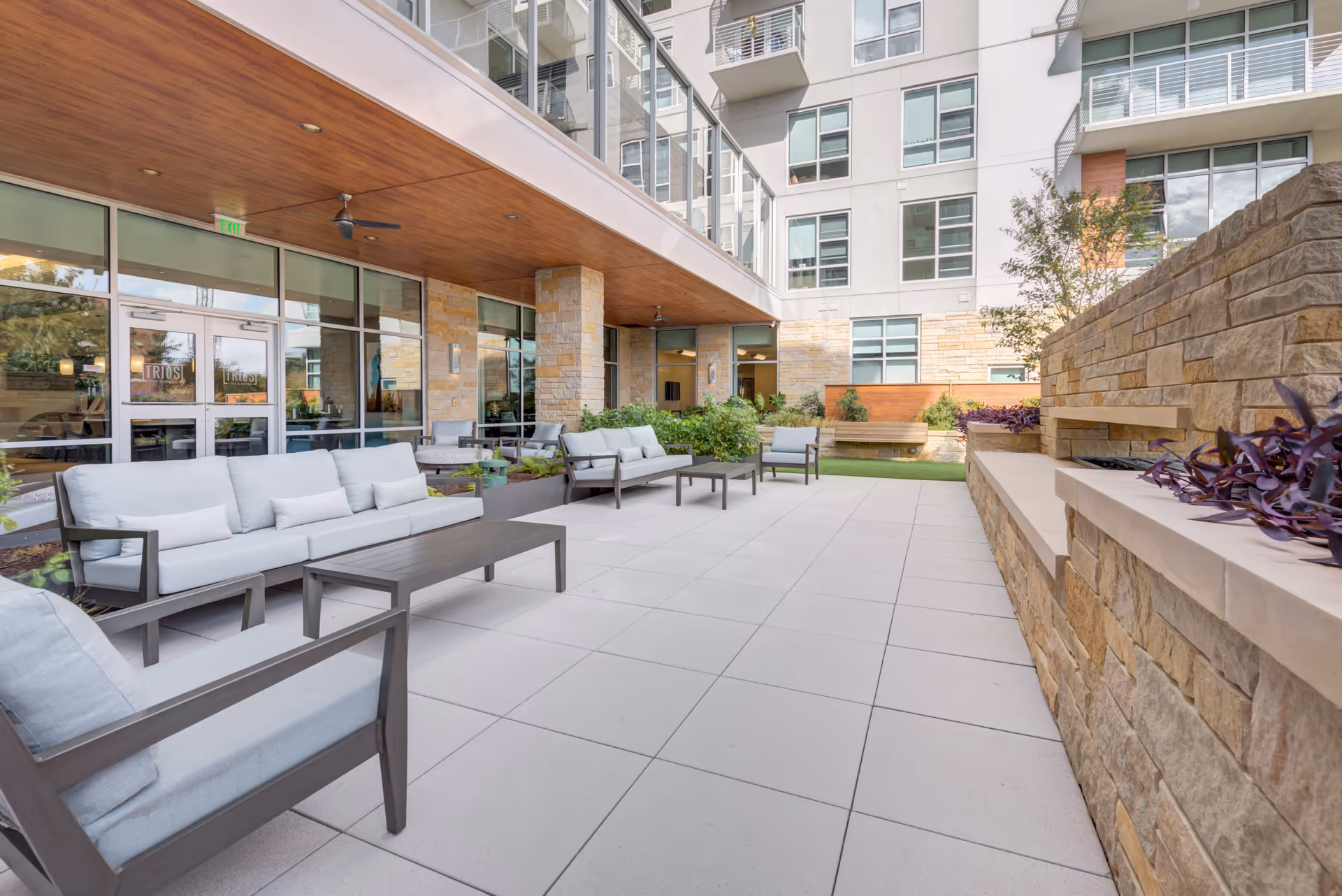 Outdoor patio area at The Village at The Triangle featuring cushioned seating with sofas and chairs, a tiled floor, stone walls, and a wooden ceiling with ceiling fans. The patio is adjacent to a building with large windows and glass doors.