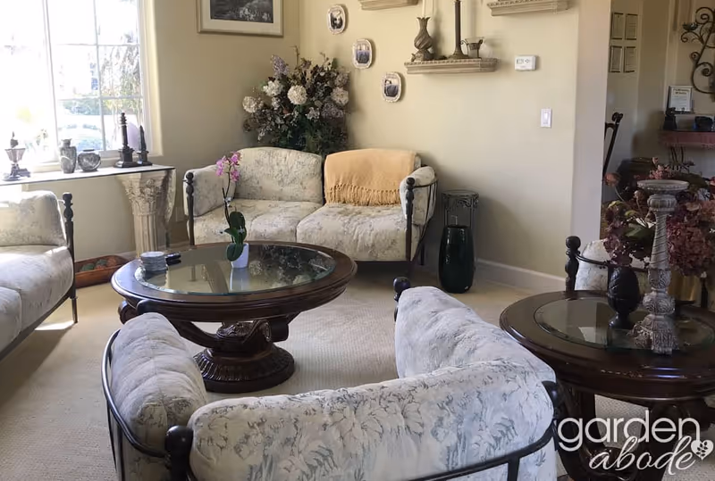 A cozy living room with floral-patterned cushioned sofas arranged around two round glass-top wooden coffee tables. The room has a large window letting in natural light, decorative shelves with small sculptures and plates on the wall, and floral arrangements adding a touch of greenery.