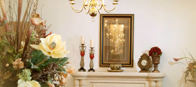 A decorative interior scene featuring a white mantel with ornate candlesticks, a small clock, a vase with red flowers, and a framed mirror reflecting a chandelier. There are also floral arrangements on both sides of the mantel.