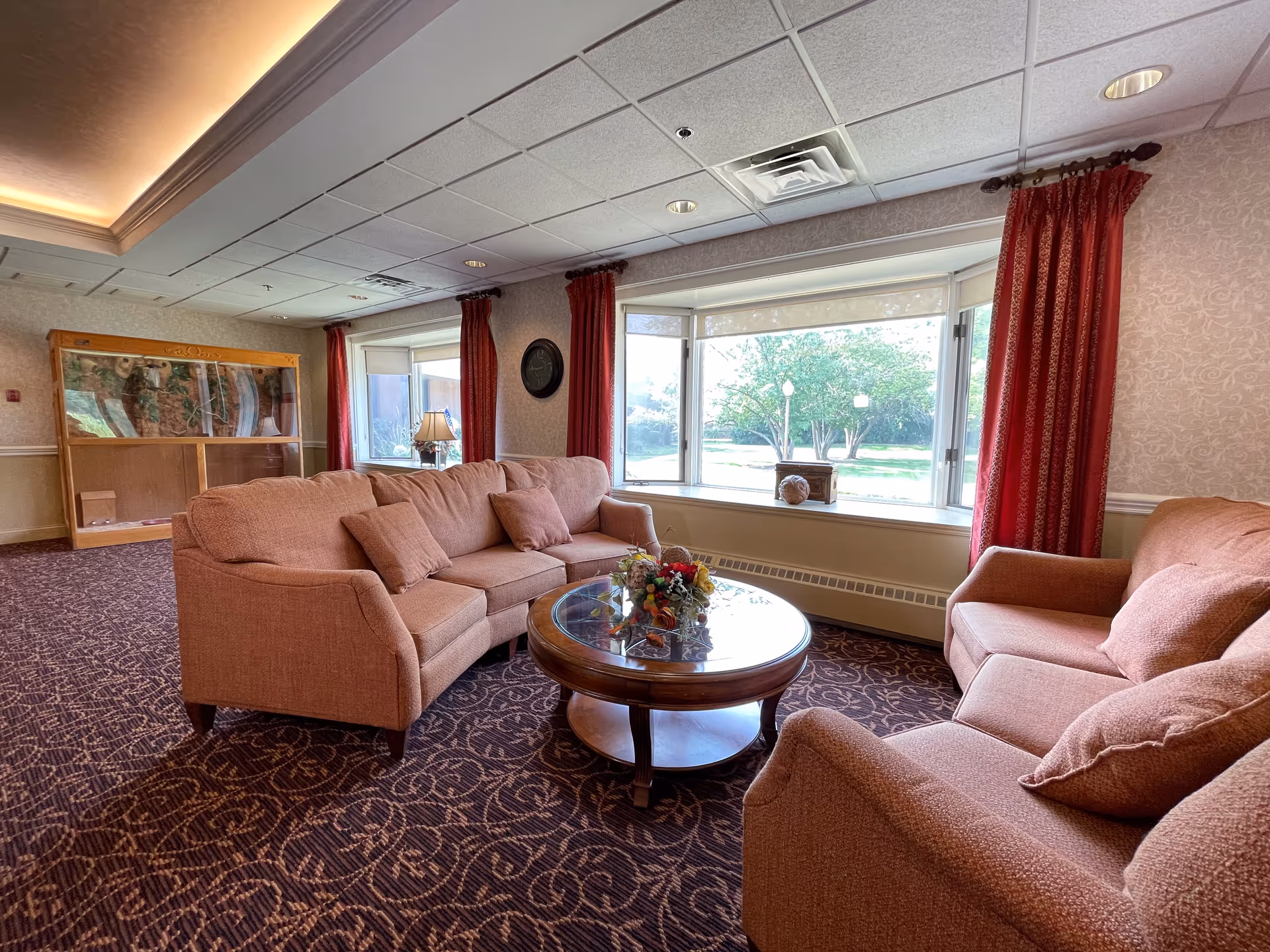 A cozy living room area with two peach-colored sofas and a round wooden coffee table with a floral centerpiece. The room has large windows with red curtains, allowing natural light to brighten the space. The carpet has a dark pattern, and there is a display cabinet and a lamp in the background.