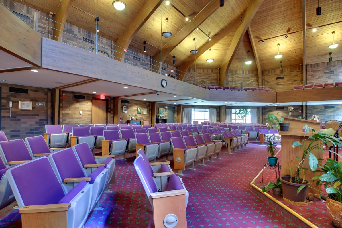 Interior view of a large auditorium or meeting room with rows of purple cushioned seats, a wooden podium with potted plants, a high vaulted wooden ceiling with hanging lights, and brick walls. There is a balcony area above with additional seating and a clock on the wall.