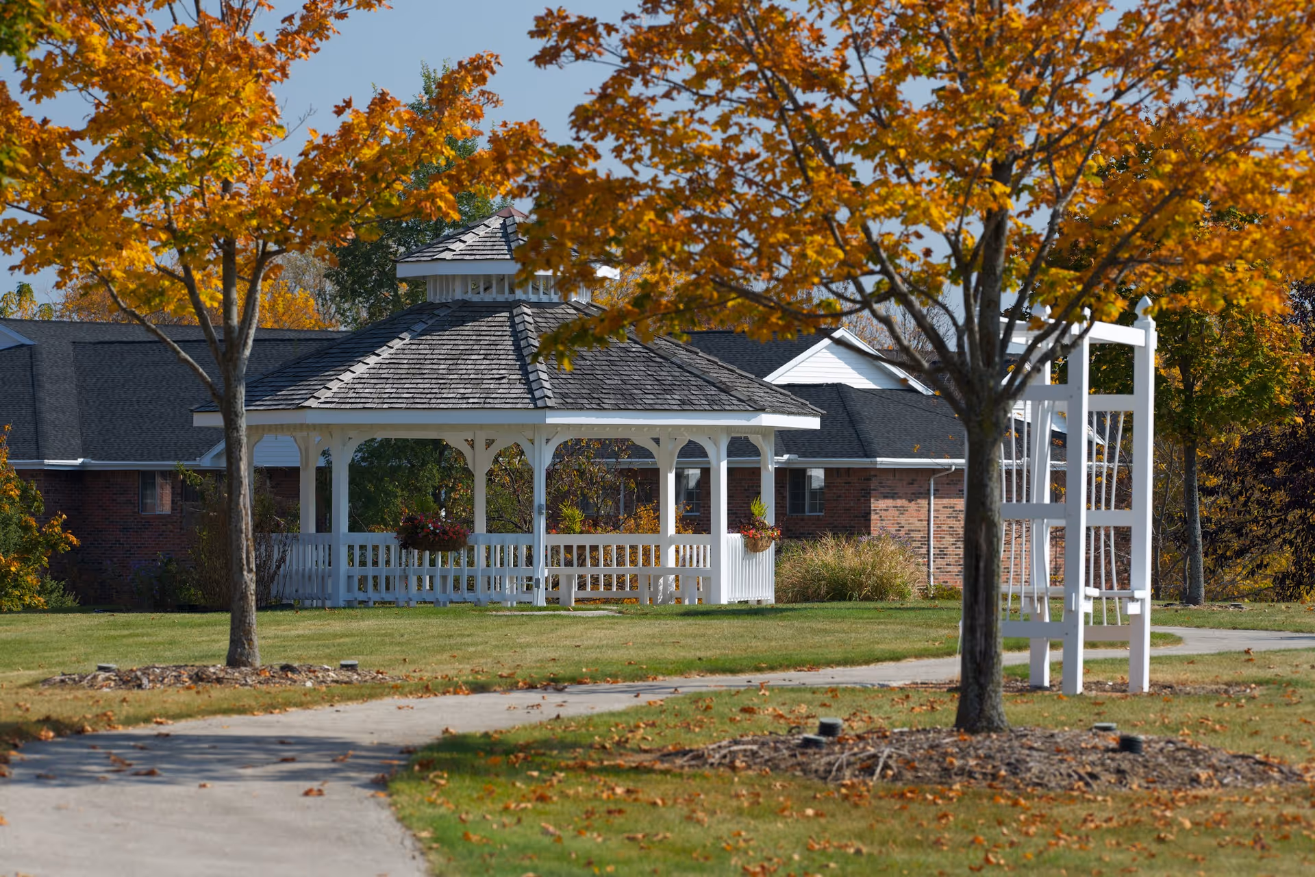 A white wooden gazebo with a shingled roof is situated on a grassy area surrounded by trees with autumn-colored leaves. A paved walkway curves through the lawn leading towards the gazebo. In the background, there is a brick building with a dark roof partially visible.