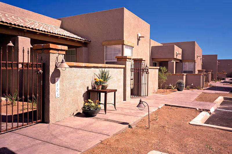 Exterior view of a row of single-story residential units with stucco walls and tiled roofs under a clear blue sky. Each unit has a small gated patio area with potted plants and outdoor lighting. A paved walkway runs in front of the units alongside a parking area.