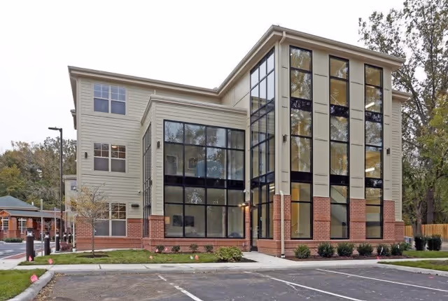 Exterior view of a modern three-story building with large vertical windows and a combination of beige siding and red brick accents, surrounded by a parking lot and some greenery.