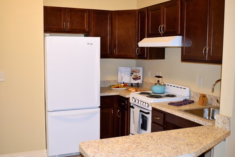 A compact kitchen with dark wooden cabinets, a white refrigerator, a white electric stove with a teal kettle on top, a beige countertop with a sink, and a cookbook stand displaying an open cookbook. There is also a plate with fruit and pastries on the counter.