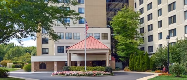 Entrance of a multi-story senior living facility with a covered drop-off area featuring a red roof. There is a flagpole with an American flag in front, surrounded by a circular flower bed. The building is surrounded by trees, shrubs, and well-maintained landscaping.
