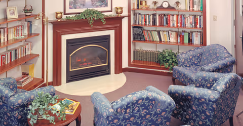 Cozy living room area with three blue floral upholstered armchairs arranged around a small wooden coffee table with a plant and books. The room features a fireplace with a wooden mantel decorated with plants and small vases. Surrounding the fireplace are built-in bookshelves filled with books and decorative items. A framed picture hangs above the fireplace.