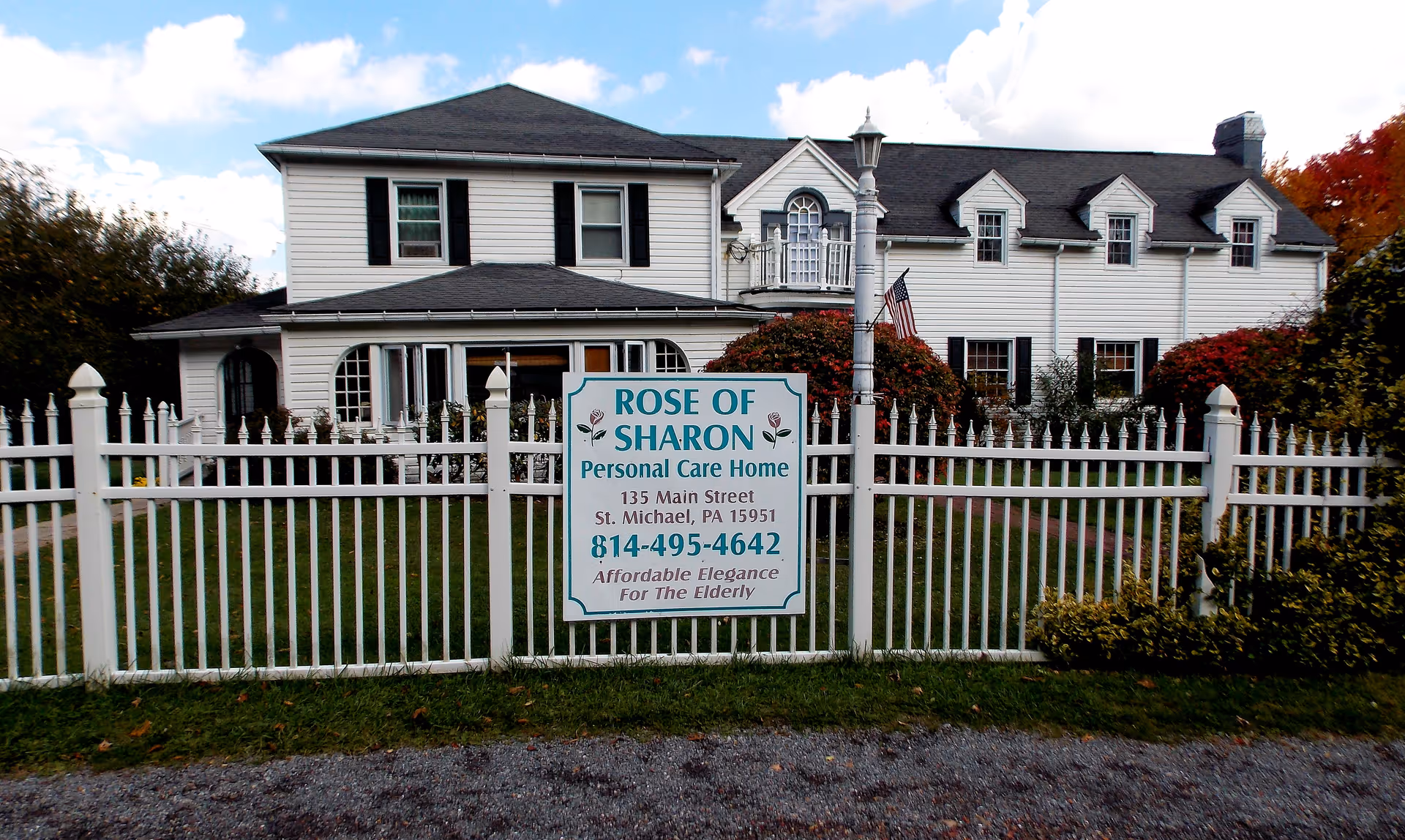 White two-story house with black shutters behind a white picket fence. A sign on the fence reads 'Rose of Sharon Personal Care Home, 135 Main Street, St. Michael, PA 15951, 814-495-4642, Affordable Elegance For The Elderly'. There is a lamppost and some bushes near the fence, and the sky is partly cloudy.