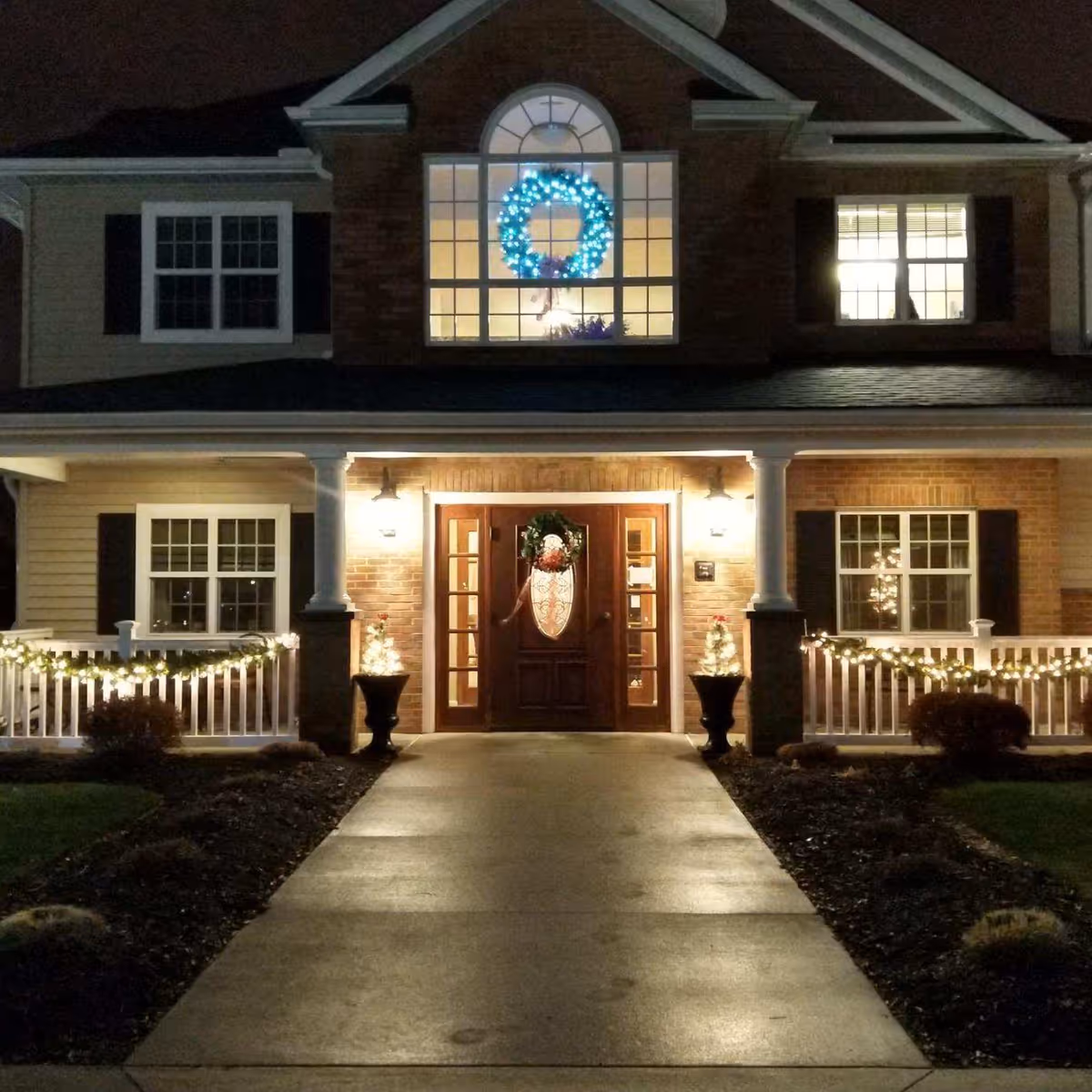 Front entrance of a two-story building at night decorated with holiday lights and wreaths. The pathway leading to the door is illuminated, and the porch railing is adorned with string lights. A large blue-lit wreath is visible in the upper window above the door.