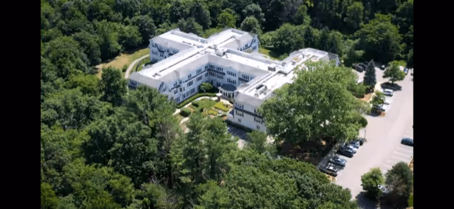 Aerial view of a large, white senior living facility building surrounded by dense green trees and vegetation, with a parking lot visible on the right side.