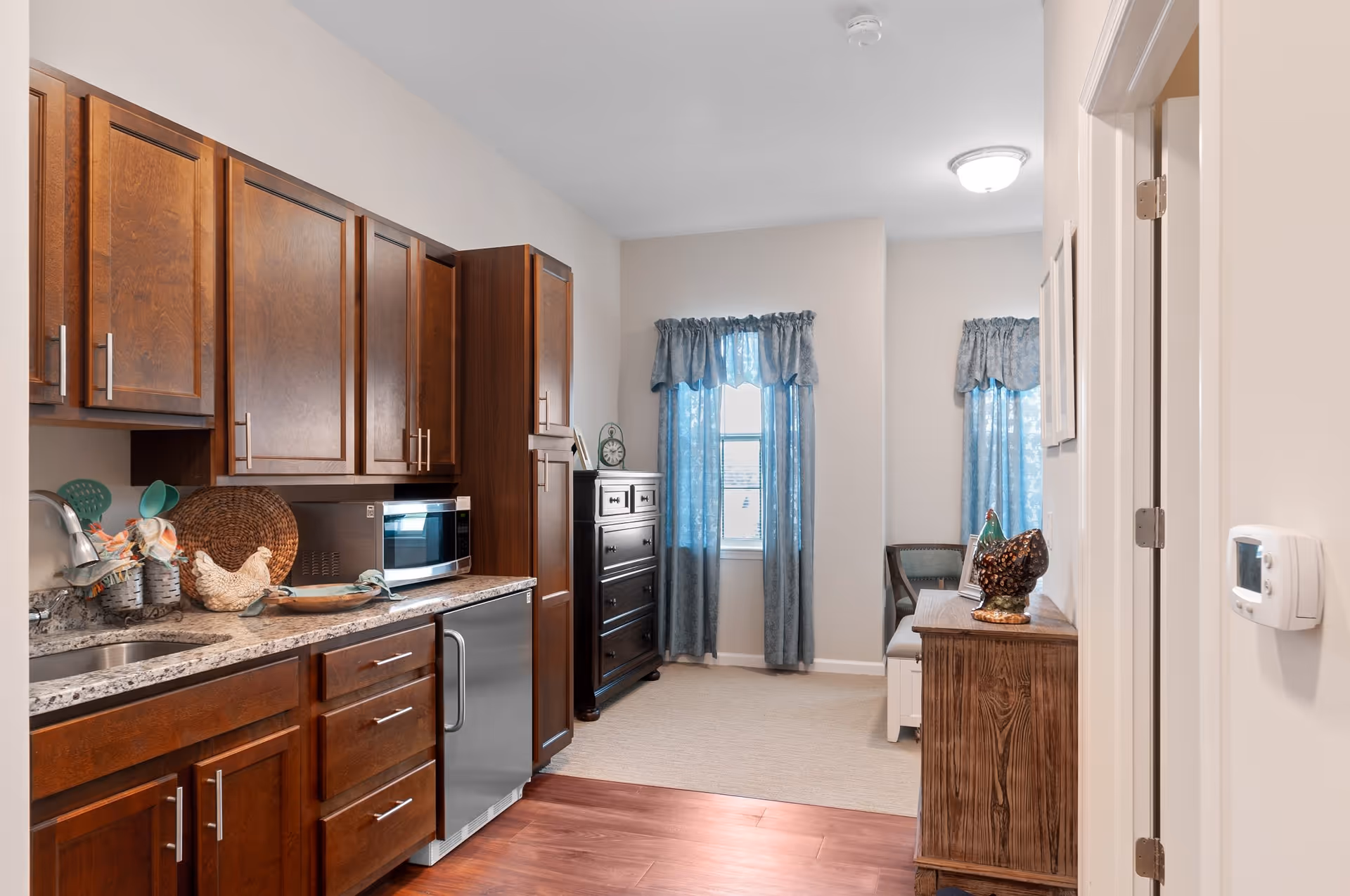 Interior view of a senior living facility kitchen area with dark wood cabinets, a granite countertop, a microwave, and a small dishwasher. Adjacent to the kitchen is a small sitting area with a wooden dresser, a chair, and windows with blue curtains letting in natural light.