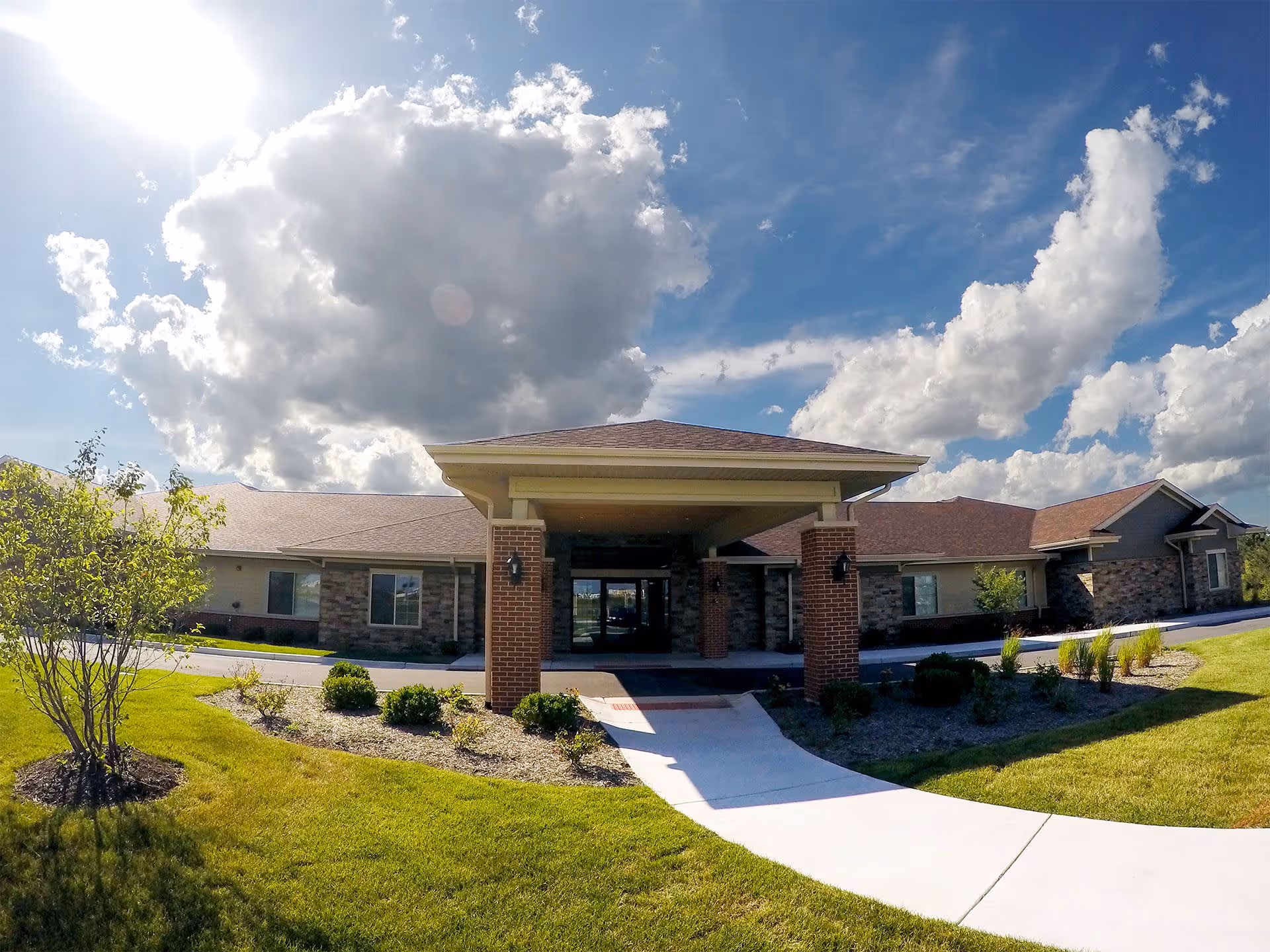 Front exterior view of AVIVA Merrillville facility with a covered entrance, brick pillars, well-maintained landscaping, green grass, and a partly cloudy blue sky.