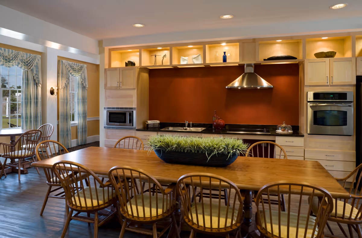 A spacious kitchen and dining area with a large wooden table surrounded by wooden chairs with yellow cushions. The kitchen features light wood cabinetry, a stainless steel oven, microwave, and range hood, with a dark countertop and a red backsplash. There are decorative items on the upper shelves and a long planter with green plants on the dining table. Two windows with patterned curtains allow natural light into the room.