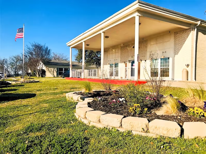 Exterior view of a single-story brick building with a covered porch supported by white columns. In front of the building is a landscaped garden bed with flowers and shrubs, bordered by stone blocks. An American flag on a flagpole is visible on the left side, with a clear blue sky overhead.